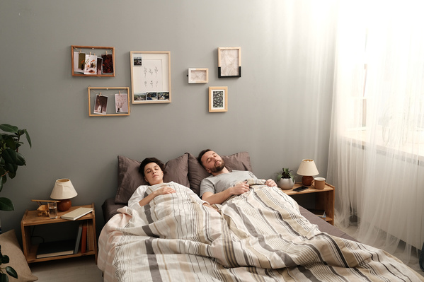 Dark-haired husband and wife in light pajamas sleeping under a white blanket in a bright bedroom