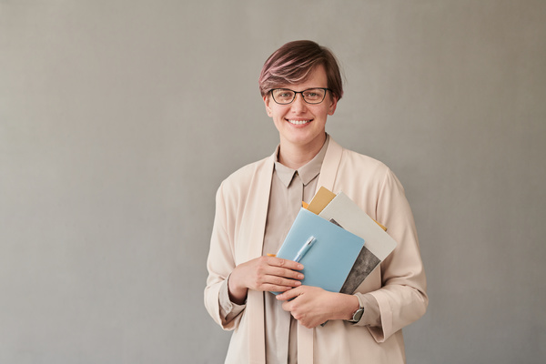 Portrait of a smiling teacher with short hair wearing glasses and strict light-colored clothes holding notebooks in her hands