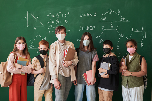 A beautiful class teacher dressed in an office suit and her smiling students with textbooks with medical masks on their faces against the blackboard