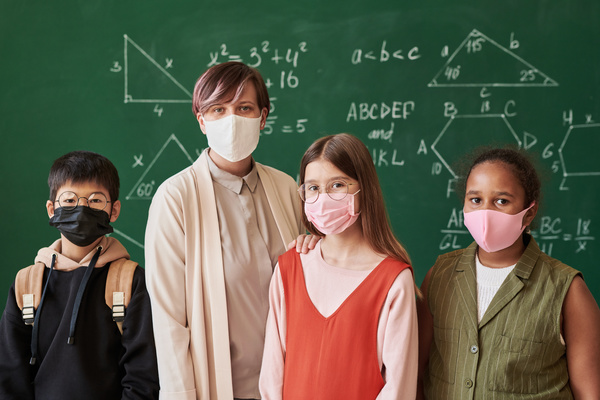 A beautiful teacher dressed in an office suit and her smiling students with textbooks in face masks against the blackboard