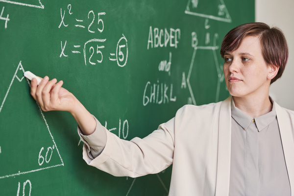 A teacher with short hair dressed in a light-colored outfit leading a geometry lesson at a chalkboard