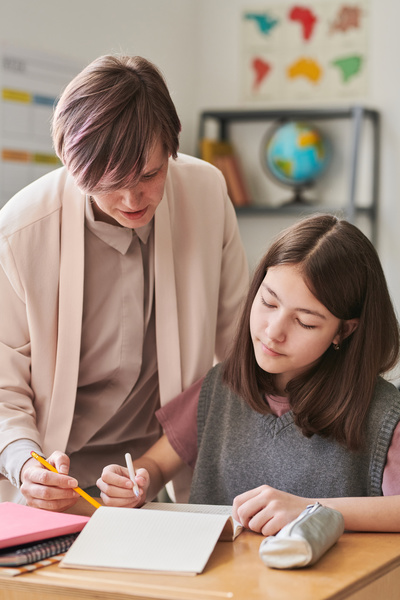 A teacher helping a student sitting at a school desk in performing written educational tasks