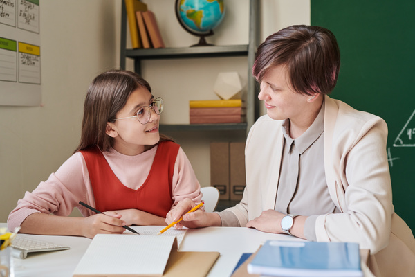 A teacher with short hair explaining a schoolgirl with glasses educational material sitting at a desk in the classroom