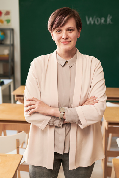 A young teacher with short hair wearing a beige suit standing in the classroom with her arms crossed on her chest