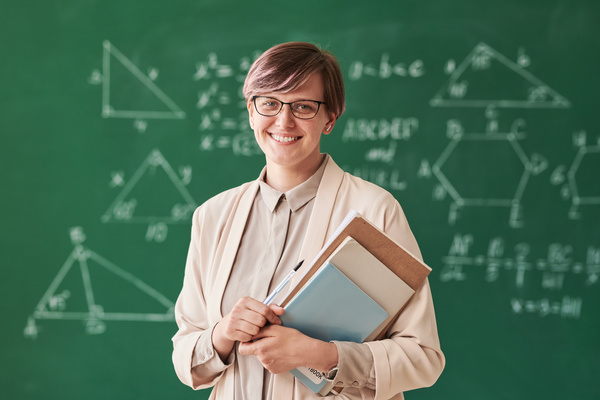 A smiling tutor with short hair wearing glasses and a beige suit holding notebooks in class