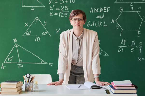 A teacher with short hair wearing glasses and a beige suit leaning on a desk in a classroom