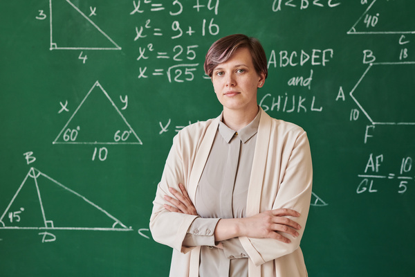 A beautiful teacher with short hair wearing a beige suit standing against a chalkboard with her arms crossed on her chest