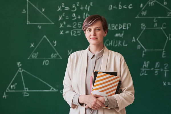A beautiful teacher with short hair wearing a beige suit holding notebooks against a chalkboard