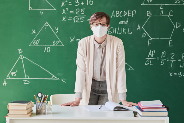 A teacher with short hair in a face mask and a beige suit leaning on a desk in the classroom