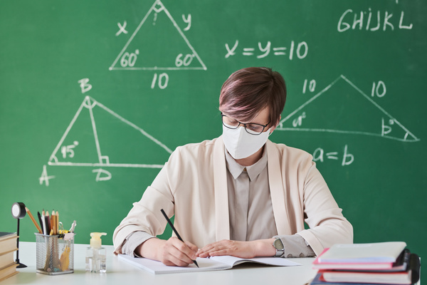 A teacher with short hair in a face mask and a light suit working at a desk in the classroom