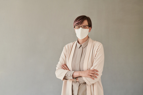 A beautiful teacher in black glasses and a face mask dressed in a light-colored office suit with her arms crossed over her chest