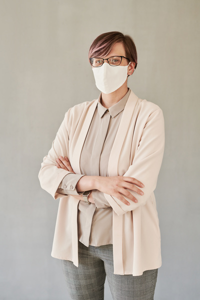 A beautiful teacher wearing a medical mask and a light-colored office suit standing with her arms crossed over her chest