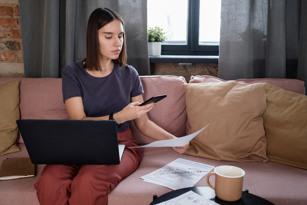 A woman with short brown hair in dark-colored clothes taking photos of work papers while sitting on the couch with a laptop on her lap