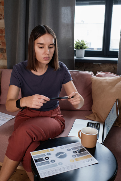 A woman with short brown hair in dark-colored clothes making photos of work papers while sitting on the couch with a laptop