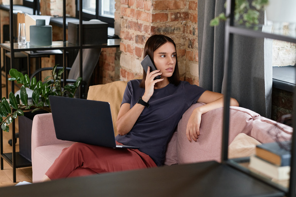 A female remote employee with short dark hair talking on the phone while sitting on the couch with a laptop on her lap