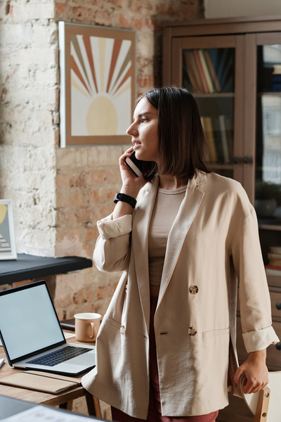 A woman with dark short hair in a beige jacket talking on the smartphone in a loft-style office
