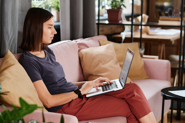 A remote female company employee with short dark hair working on a laptop while sitting on a pink sofa