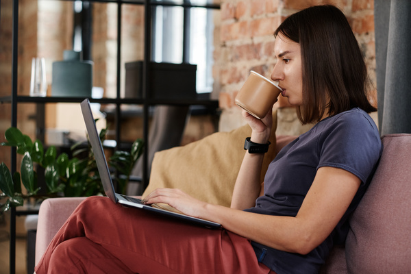 A woman with short dark hair working on a laptop while sitting on a pink sofa and drinking tea