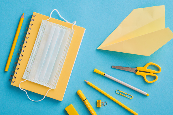 Top view of a yellow stationery set with a medical mask and a yellow paper airplane on a blue surface