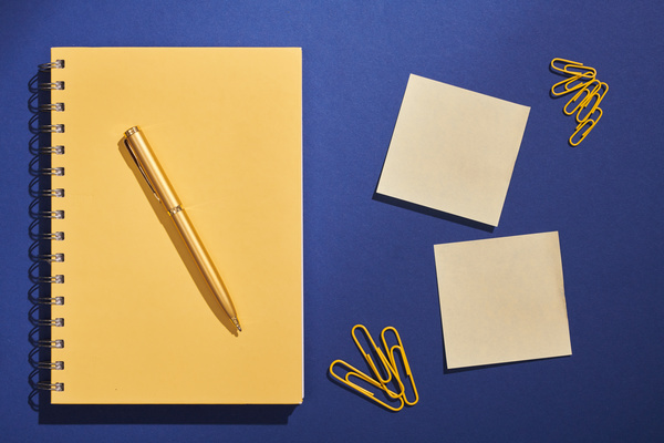 A yellow spiral notebook with a gold pen on it and stationery laid out on a dark blue surface
