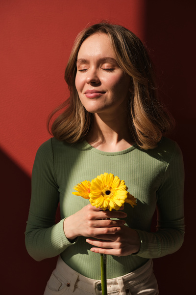 A blonde woman with closed eyes dressed in green longsleeve smiling tenderly and holding yellow flowers