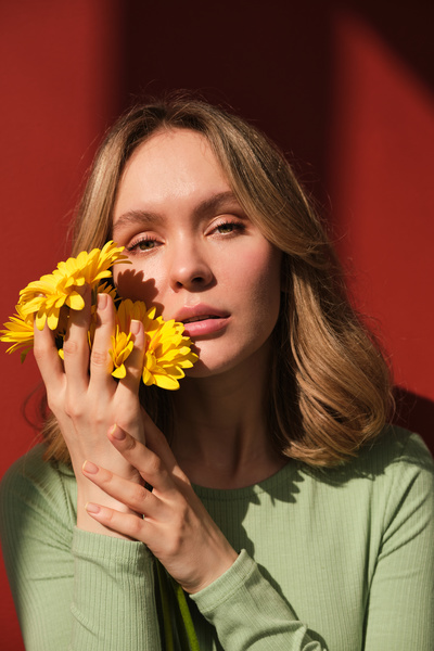 A blonde woman dressed in green longsleeve cuddling up yellow gerberas to her face against a red background