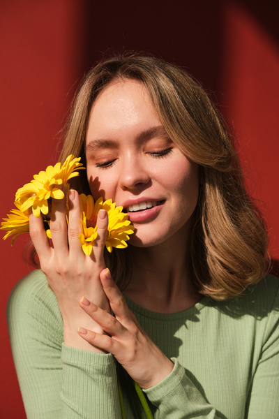A woman dressed in green longsleeve with closed eyes cuddling up yellow gerberas to her face against a red background