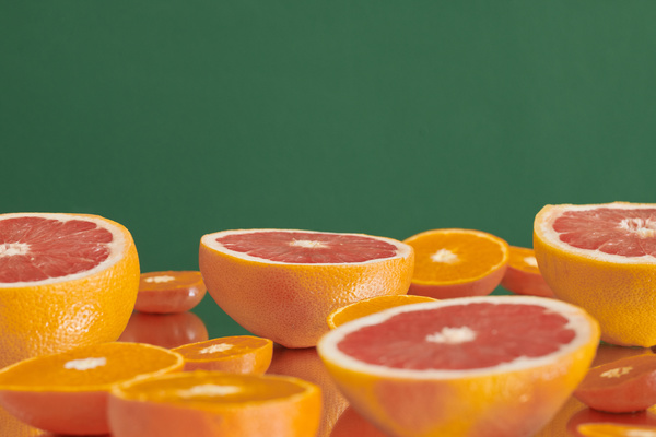 Halves of grapefruits and oranges are chaotically laid out on a reflective surface against a green background