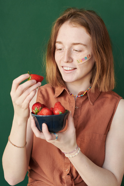 A young woman with red hair and freckles looking at a strawberry taken from a black bowl