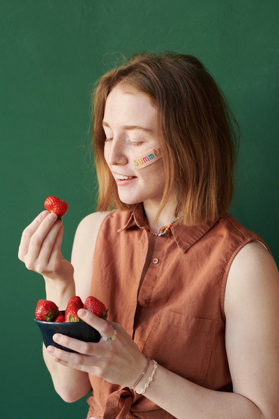 A young woman with red hair and freckles eating a strawberry from a black bowl