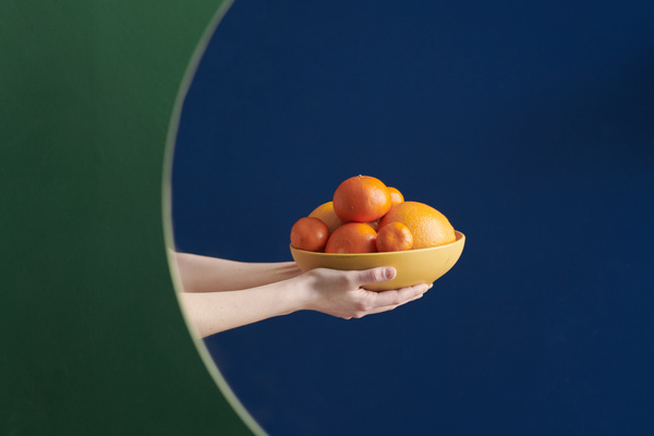 Reflection of a yellow bowl with citrus fruits in hands in a mirror hanging on a green wall