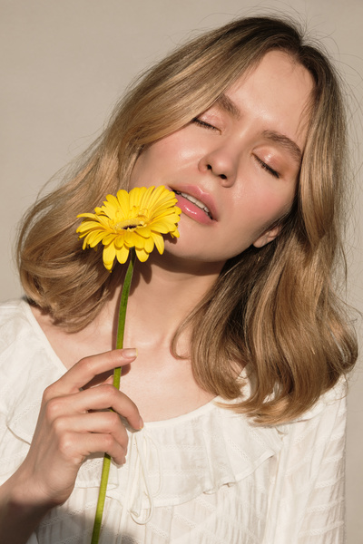 A blonde woman with closed eyes dressed in white clothes poses with a yellow gerbera