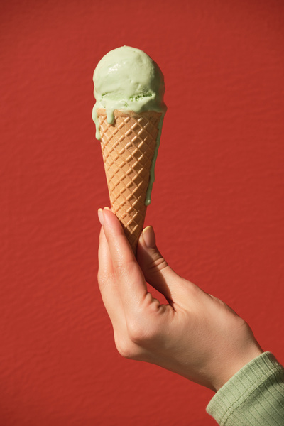 Melted pistachio-flavored ice cream in a cone is held in the left hand against a red background