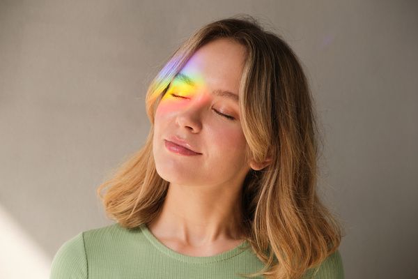 A woman with short blonde hair and a rainbow ray on her face closed her eyes against a white background