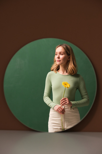 Mirror reflection of a woman in light clothes with light short hair holding a yellow gerbera