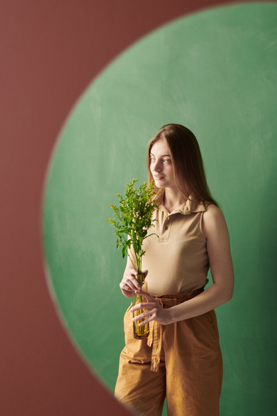 Reflection in the mirror hanging on the red wall of a woman with long blond hair in beige clothes holding a vase with a sprig of wild flowers