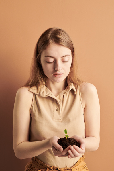 A woman with long brown hair dressed in a beige outfit carefully holding a handful of earth with a sprout