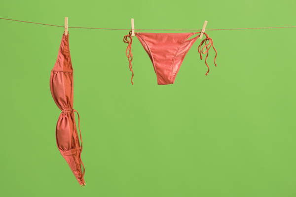 Coral womens swimsuit attached with clothespins hanging on a drying rope against a green background