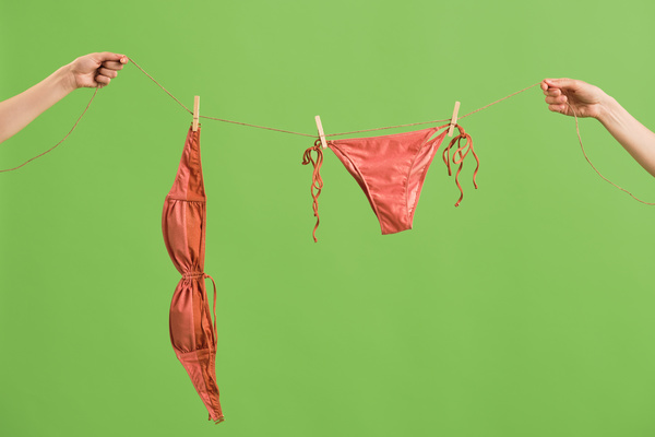 A coral female bikini attached with clothespins hanging on a drying rope which is in the hands against a green background