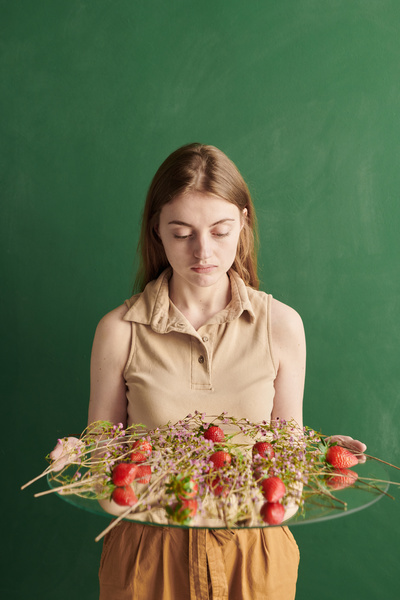 A woman with long brown hair looking at a composition of pink wildflowers and strawberries on a round mirror