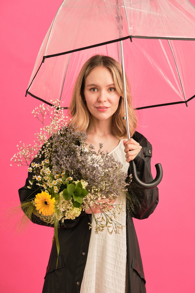 A woman with blonde hair in a black raincoat holding a transparent umbrella and a bouquet of flowers