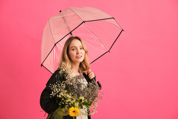 A woman with blonde hair in a black raincoat posing with a transparent umbrella and a bouquet of flowers