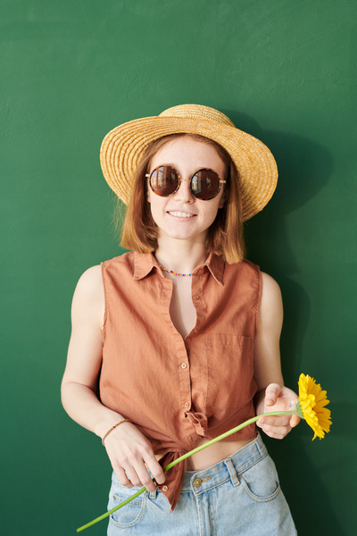 A woman with red hair in a summer straw hat and sunglasses with a yellow gerbera at her face