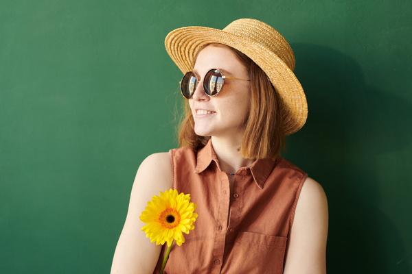 A woman with red hair wearing a summer straw hat and round sunglasses holding a yellow gerbera