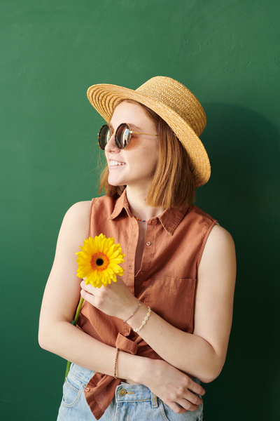 A red-haired woman wearing a straw hat and glasses with yellow gerbera in her hands against bright background