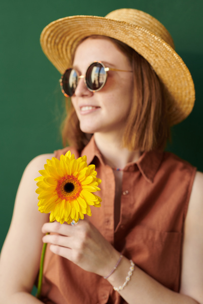 A smiling woman in a summer outfit with a straw hat and sunglasses holds a bright gerbera against a green background