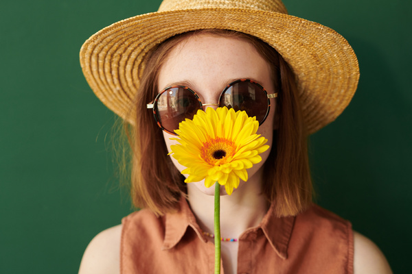 A woman in summer clothes with a straw hat on her head and sunglasses smiles holding a bright gerbera