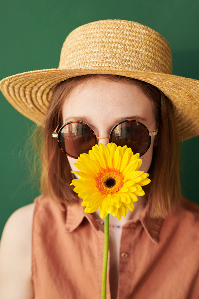 A woman in summer clothes with a straw hat on her head and sunglasses posing with a yellow gerbera