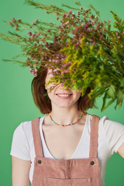 A smiling woman with short red hair dressed in summer clothes with a colorful beaded necklace hiding her eyes with a bouquet of wildflowers