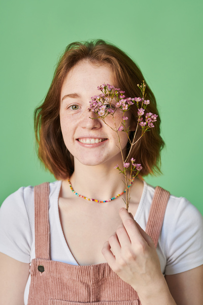 A smiling woman with a red bob and a colored beaded checker around her neck holding pink wildflowers are in front of her face